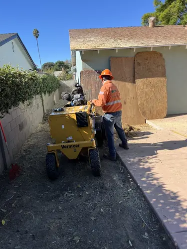 Worker grinding a tree stump with heavy equipment beside a house